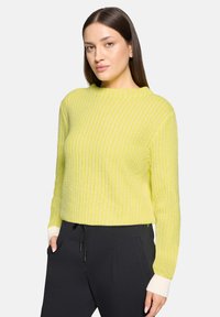 Frau mit langen braunen Haaren, die einen limettengrünen, gerippten Pullover mit weißen Bündchen und schwarze Hose trägt, steht mit einer Hand in der Tasche.