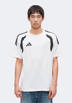 Ung mand med kort mørkt hår iført en hvid Adidas t-shirt med sorte striber på skuldrene og blå jeans, stående foran en ensfarvet baggrund.