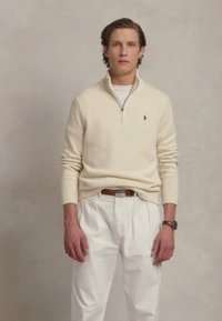 Pull en maille crème avec fermeture éclair et poignets côtelés, associé à un pantalon plissé blanc. Comprend une ceinture brune et une montre. Aucun motif ni logo visible.