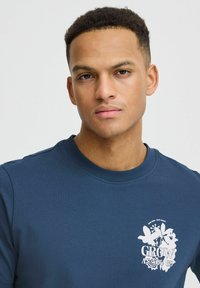 Marinblå t-shirt med kort ärm gjord av bomull och rund hals. Har en vit grafisk tryck av blommiga element och texten "VÄX EFTERHAND."