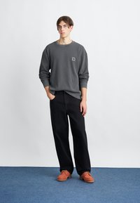 Grauer Sweatshirt mit einem Patch-Logo, schwarze, locker sitzende Hosen und orangefarbene Schnürschuhe. Der Hintergrund ist schneeweiß.