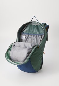 Mochila verde y azul con una gran apertura, forro interior gris y múltiples compartimentos. Presenta un asa superior y bolsillos laterales.