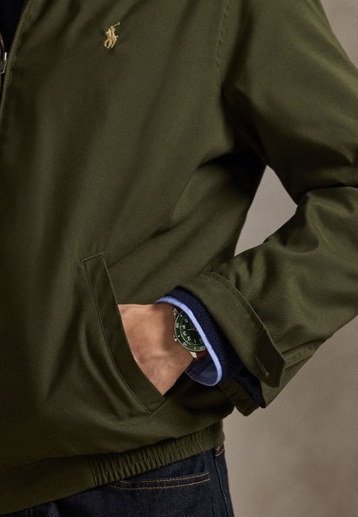 Temno zelena jakna z zadrgo in rebrastimi manšetami, z žepom in izvezenim logotipom. Zapestna ura ima zelen obraz, srebrne poudarke in črn pašček.