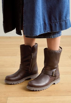 Brązowe skórzane botki do kolan z bocznym zamkiem, detalem w postaci paska i teksturowaną podeszwą, umieszczone na jasnym drewnianym podłodze.