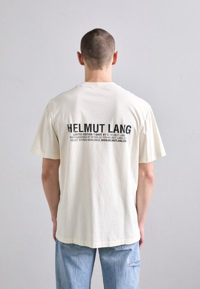 Kreemvärvi puuvillane T-särk lühikeste varrukatega, mille taga on julge must kiri, mis sisaldab brändi "HELMUT LANG" ja muid detaile.
