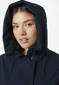 Chaqueta impermeable de color azul marino con un alto cuello, capucha fija y cremallera frontal. Presenta una textura suave y cordones ajustables.