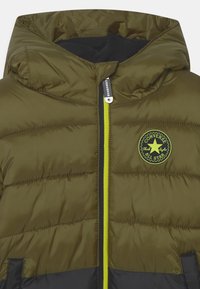Giubbotto imbottito di colore verde oliva con fondo nero, dotato di chiusura con zip, cappuccio e zip con accentuazione gialla. Include un logo rotondo di Converse.