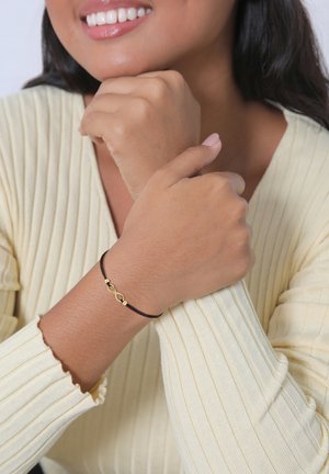 Bracciale di corda nera con un simbolo di infinito dorato e due perle dorate. Indossato su un polso, con una maglietta a maniche lunghe in giallo chiaro a coste.