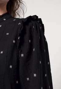 Blusa preta com padrões brancos em relevo, apresentando detalhes de ombros com babados e um tecido texturizado, destacando os seus elementos de design únicos.