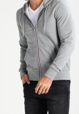 Hombre con sudadera con capucha gris claro y cremallera, con cordones blancos, sobre una camiseta blanca y vaqueros negros, con las manos cerca de los bolsillos delanteros.
