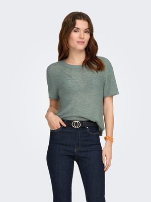 Femme portant un haut vert en maille à manches courtes rentré dans un jean bleu foncé avec une ceinture noire et un bracelet en perles orange, debout devant un fond uni.