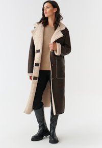 Manteau en simili cuir marron foncé, long, avec un col en peluche beige, comportant cinq boutons et des poches latérales. Porté par-dessus un haut beige et un pantalon noir.