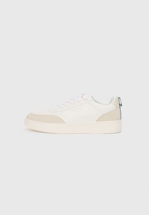 Sneaker blanche avec des accents en suède beige, lacets plats et semelle en caoutchouc texturé. Inclut un détail de boucle verte au niveau du talon.