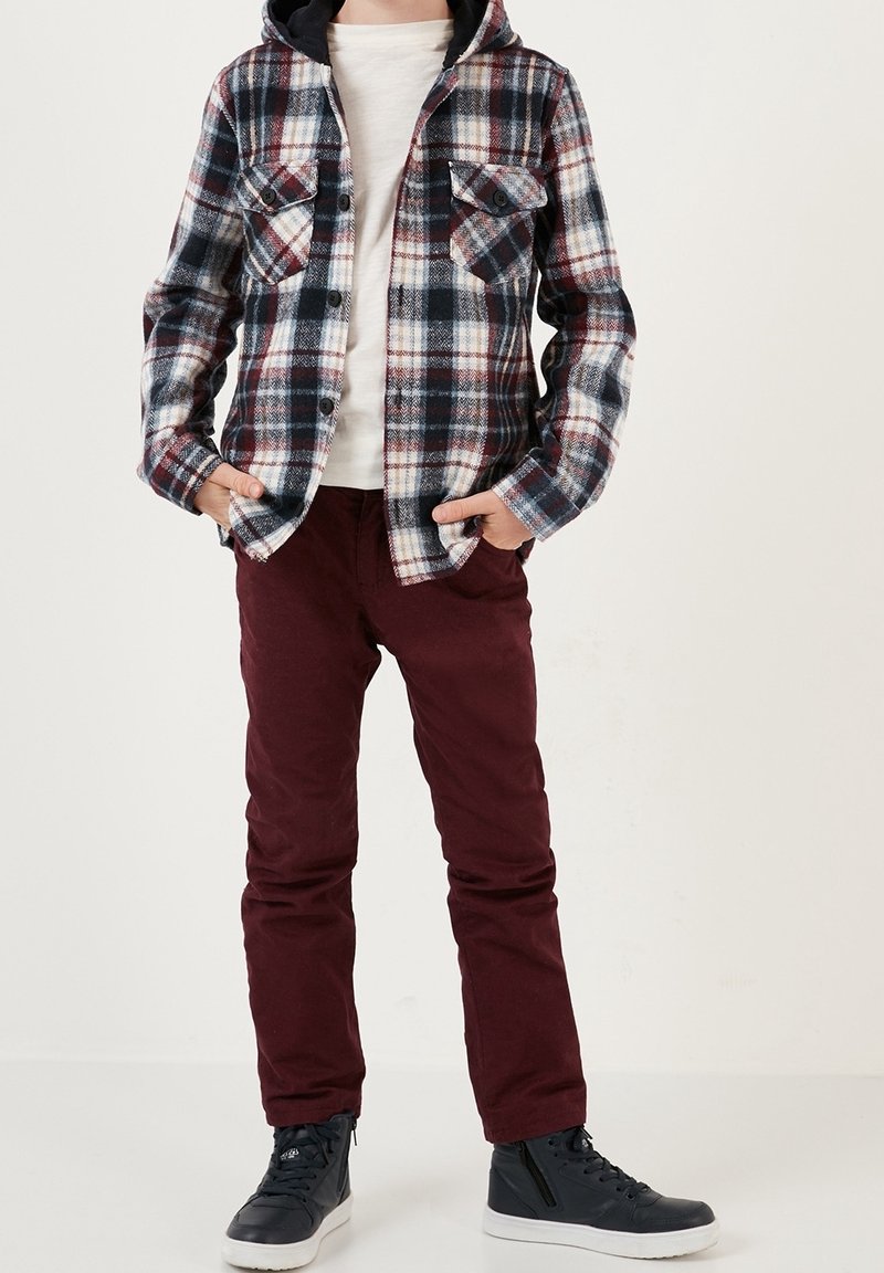 Chemise en flanelle à carreaux bleu, blanc et bordeaux, avec des boutons et deux poches avant. Associée à un pantalon bordeaux et des baskets montantes noires.