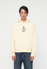 Sudadera amarilla con bolsillo frontal, que presenta un logo negro de "tommy girl" y un acento de estrella. Combinada con jeans de mezclilla oscura.