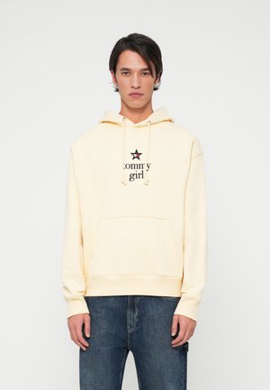 Sudadera amarilla con bolsillo frontal, que presenta un logo negro de "tommy girl" y un acento de estrella. Combinada con jeans de mezclilla oscura.