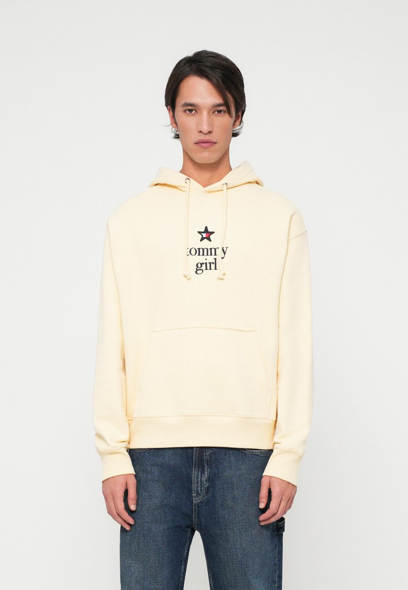 Sudadera amarilla con bolsillo frontal, que presenta un logo negro de "tommy girl" y un acento de estrella. Combinada con jeans de mezclilla oscura.