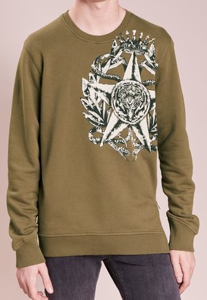 Man wearing olive green sweatshirt with black and white graphic of a star, crowned wreath, tiger face, and snake.
