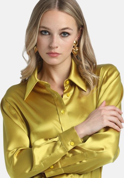 Cămașă din satin galben, cu guler ascuțit, mâneci lungi și detalii cu nasturi. Modelul poartă cercei aurii în formă de inimă. Textură netedă.