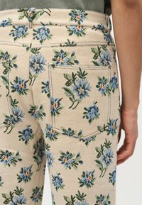 Κοντινό πλάνο μπεζ παντελονιού με μπλε και πράσινο floral μοτίβο, απεικονίζοντας την πίσω τσέπη και μέρος ενός πράσινου πουκάμισου και χεριού.