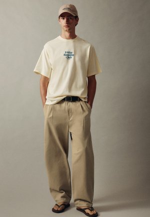 Jeune homme portant une casquette beige, un T-shirt crème avec le texte "Friday Running Club", un pantalon kaki ample, une ceinture noire et des tongs noires, debout devant un fond uni.