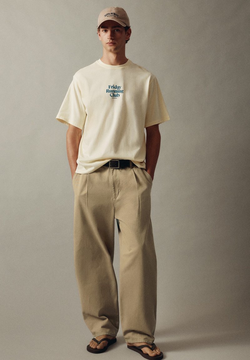 Jeune homme portant une casquette beige, un T-shirt crème avec le texte "Friday Running Club", un pantalon kaki ample, une ceinture noire et des tongs noires, debout devant un fond uni.