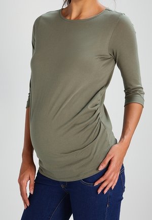 Femme enceinte portant un haut de maternité froncé vert olive à manches trois-quarts et un jean bleu foncé, debout avec une main sur la hanche.