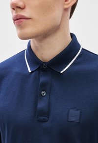 Tricou polo bleumarin cu un finisaj texturat, tiv alb contrastant la guler, nasturi dubli și un logo pătrat pe piept.