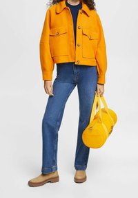 Orange cropped jacket, dark blue top, blue jeans, tan ankle boots, and a yellow duffel bag. Notable button accents on the jacket.