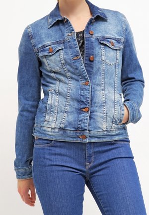 Person iført en blå denim jakke med knapper og lommer og blå jeans, den ene hånd i jakklommen, mod en ensfarvet baggrund.