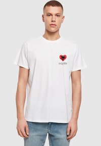 Balta medvilninė marškinė su raudonu širdies grafiku, juodu erelio dizainu ir tekstu "SHQIPËRI" po juo, trumpomis rankovėmis ir standartinio kirpimo.