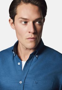 Niebieska koszula z guzikami, wykonana z teksturowanej tkaniny, z naszywaną kieszonką i subtelnym detalem logo na piersi.