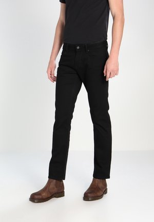 Mand iført slanke sorte jeans, sort t-shirt og brune læder ankelstøvler står på hvid baggrund.