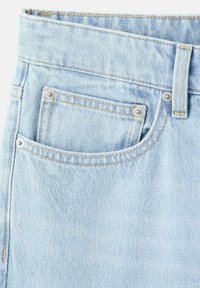 Primer plano de pantalones vaqueros de mezclilla azul claro que muestra el bolsillo delantero, el bolsillo para monedas, la trabilla del cinturón y detalles de costuras naranjas.