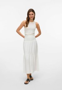 Débardeur blanc à côtes associé à une longue jupe blanche fluide avec une taille haute smockée et un design en couches. Des sandales noires complètent le look.
