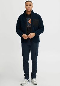 Navy fleecejack met hoge kraag en zwarte borstzak, gecombineerd met donkere cargo-broeken en grijze sneakers met blauwe accenten.