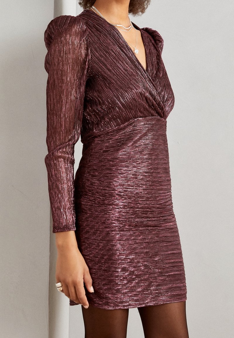 Femme portant une mini-robe texturée couleur prune brillante avec de longues manches bouffantes et des colliers argentés superposés.