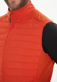 Oransje isolert vest med hög krage, quiltat mönster, framdragkedja och "ENDURANCE" tryckt på axeln. Lättviktigt tyg.
