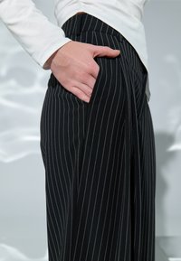 Käsi mustade triibuliste pükste taskus, vöökohal nähtav valge pika varrukaga särk.