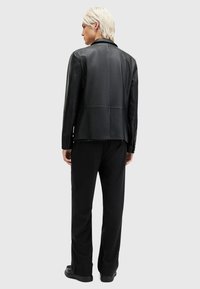 Chaqueta de cuero negro con un diseño estructurado, que presenta detalles de costura y botones en los puños, combinada con pantalones negros.