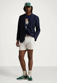 Άνδρας με πράσινο και λευκό καπέλο, μπλε ριγέ σακάκι, μπλε μπλουζάκι με γραφικό αρκούδας, λευκά σορτς, μπεζ κάλτσες και πράσινα αθλητικά παπούτσια.