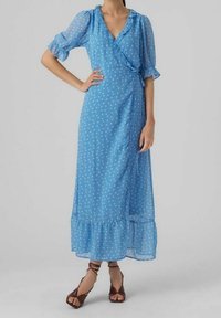 Kvinna iklädd en blå lång klänning med prickar, med volang på kragen och nederkanten, samt bruna öppna klackar med remmar.