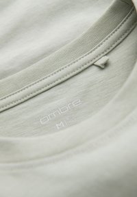 Tela de camiseta de algodón verde claro con cuello redondo, con etiqueta que muestra la marca "ombre" y la talla "M" impresa en blanco.