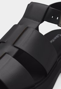 Μαύρο δερμάτινο σανδάλι με φαρδιές λουρίδες και ρυθμιζόμενο κούμπωμα γύρω από τον αστράγαλο σε παχύ πάτο.