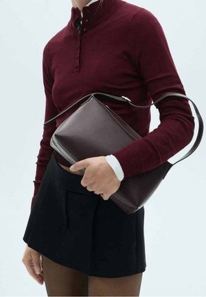 Personne portant un haut à manches longues bordeaux et une jupe noire, tenant un sac en cuir marron foncé porté en bandoulière.