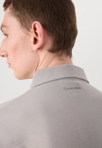 Grå skjorte med krage, vertikale striper, glatt tekstur, og diskret "Calvin Klein"-logo bak nær halsen.