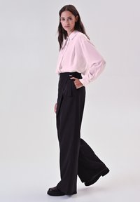 Blusa rosa chiaro a maniche lunghe con colletto lucido, abbinata a pantaloni neri a vita alta e gamba larga, con scarpe nere massicce.