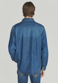 Uomo in piedi di spalle, che indossa una camicia in denim blu a maniche lunghe e jeans abbinati, con uno sfondo neutro.