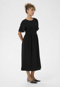 Vestido negro texturizado de longitud media con mangas cortas y cintura ceñida. Combinado con zapatos de tacón alto negro tipo slingback.