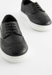 Zapatos brogue negros de cuero con detalles perforados, suela de goma blanca, punta redonda y diseño de cordones de seis agujeros. Textura suave, estilo clásico.